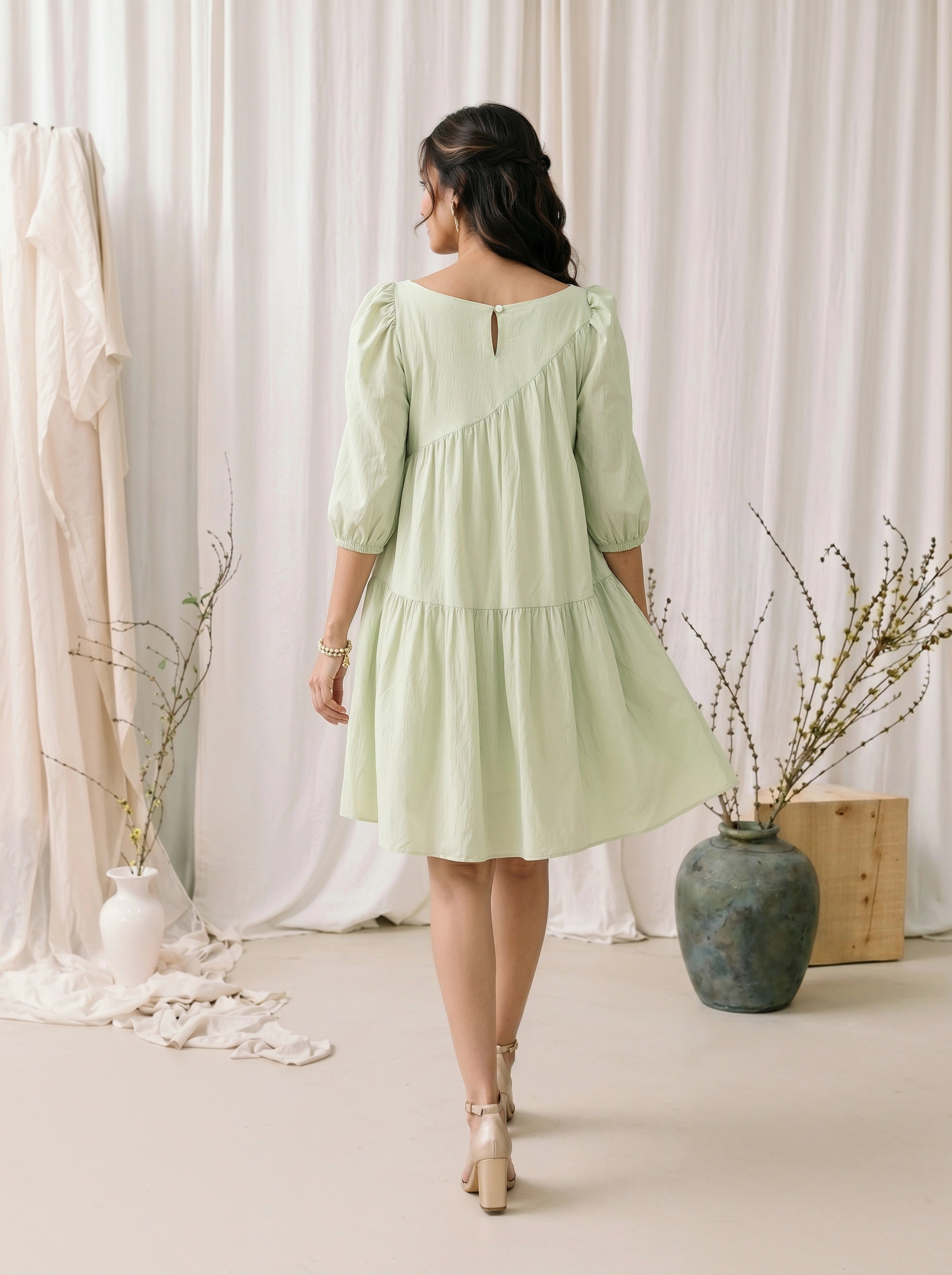 Woman in a light green dress standing in a room with white curtains and decorative vases.