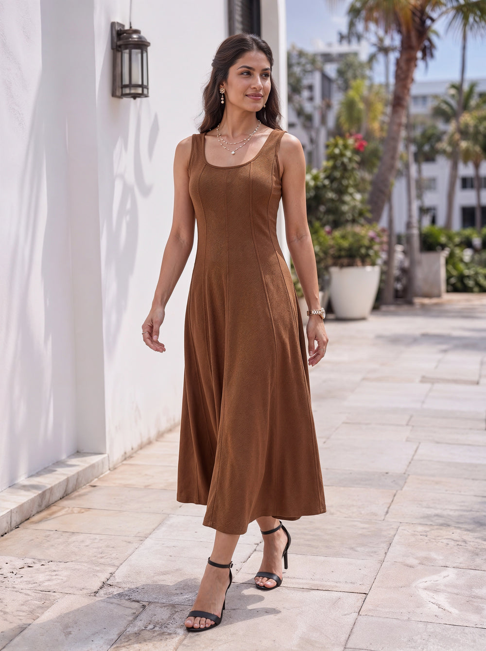 A woman wearing a sleeveless brown midi dress walks outdoors on a stone walkway next to a white wall with palm trees in the background.
