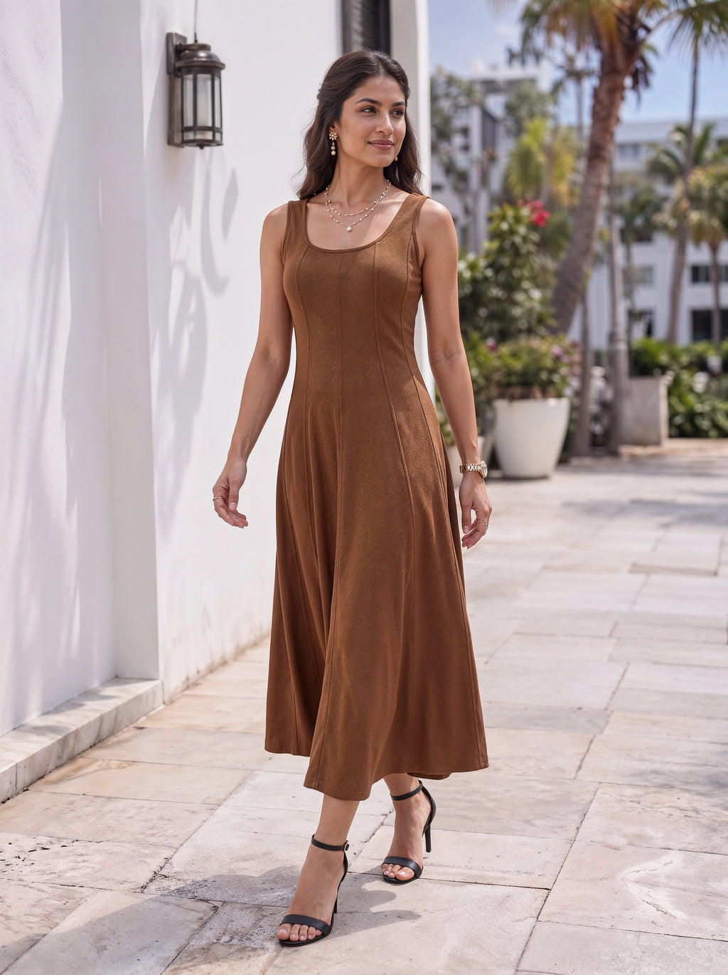 A woman wearing a sleeveless brown midi dress walks outdoors on a stone walkway next to a white wall with palm trees in the background.