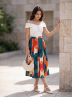 A woman stands outdoors against a stone wall wearing an off-the-shoulder white top and a midi skirt with a bold abstract geometric pattern in teal, orange, cream, and burgundy. She holds a cream handbag and wears white heeled sandals.