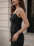 A woman wearing a black lace-up spaghetti strap camisole top and matching black trousers, standing outdoors against a light beige stone building wall.