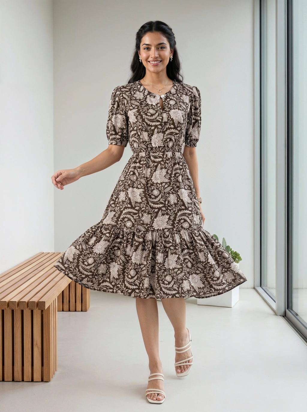 A woman modeling a short-sleeve midi dress with a brown and beige floral toile pattern, standing in a bright, modern room with a wooden bench and large windows.