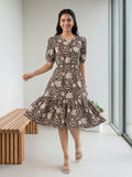 A woman modeling a short-sleeve midi dress with a brown and beige floral toile pattern, standing in a bright, modern room with a wooden bench and large windows.