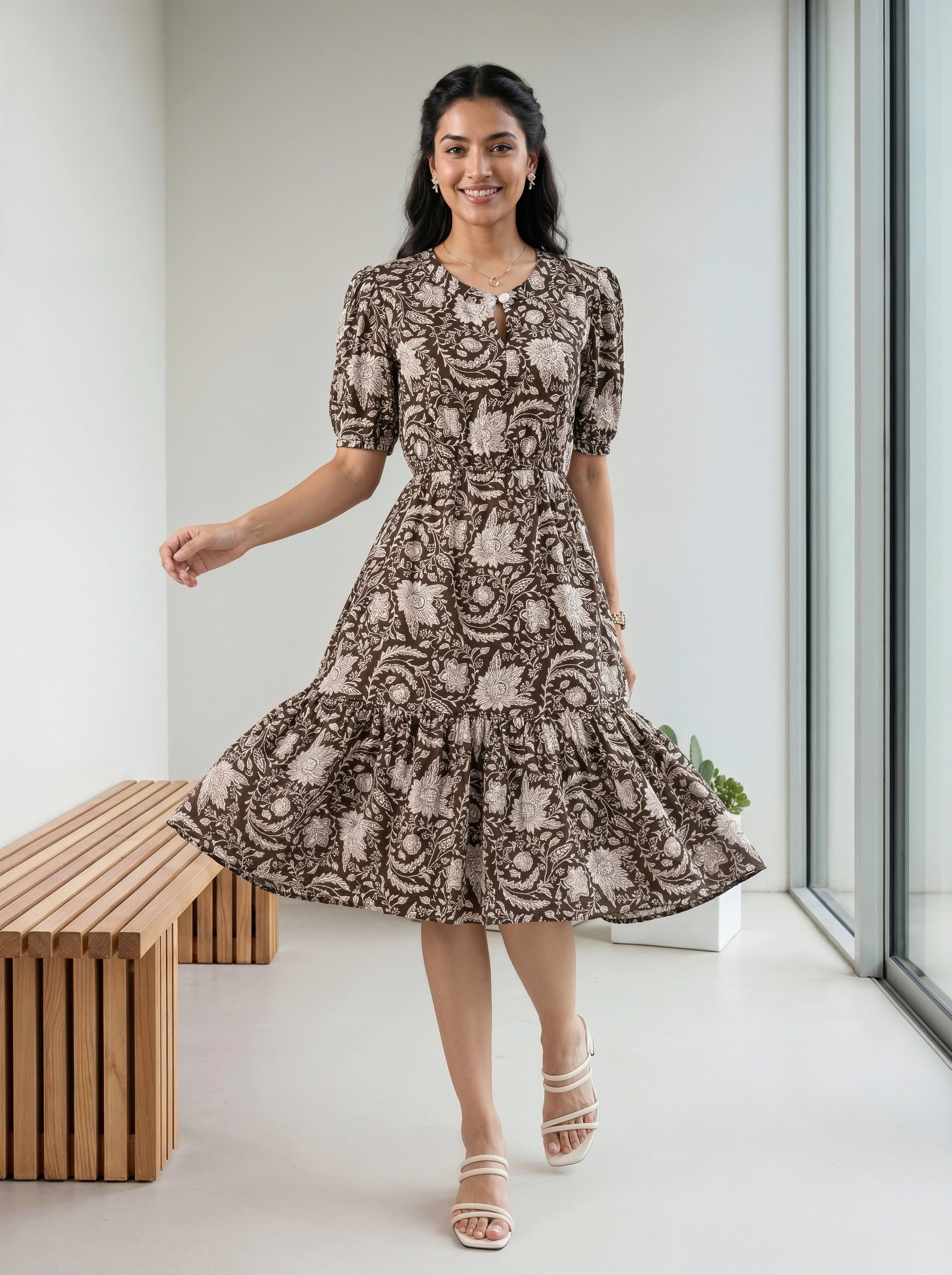 A woman modeling a short-sleeve midi dress with a brown and beige floral toile pattern, standing in a bright, modern room with a wooden bench and large windows.