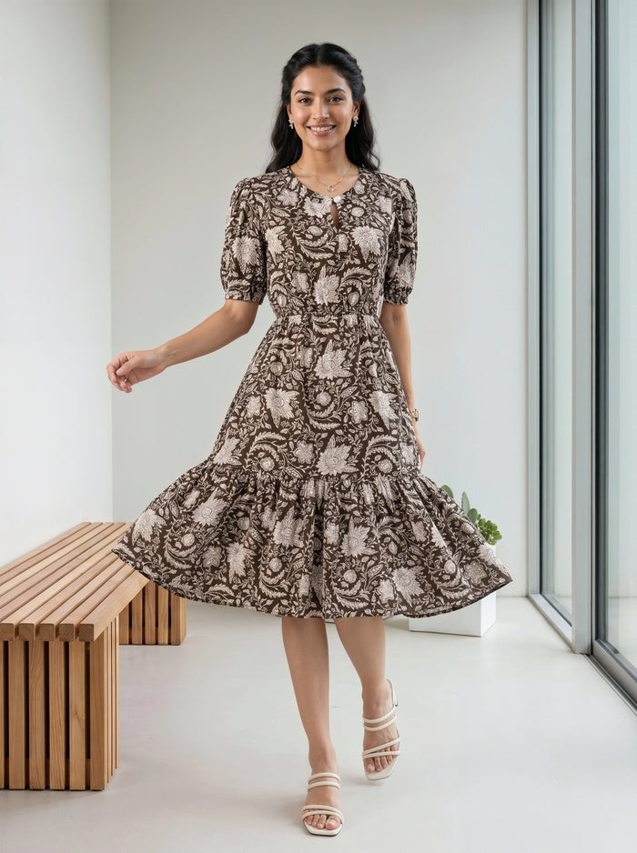A woman modeling a short-sleeve midi dress with a brown and beige floral toile pattern, standing in a bright, modern room with a wooden bench and large windows.