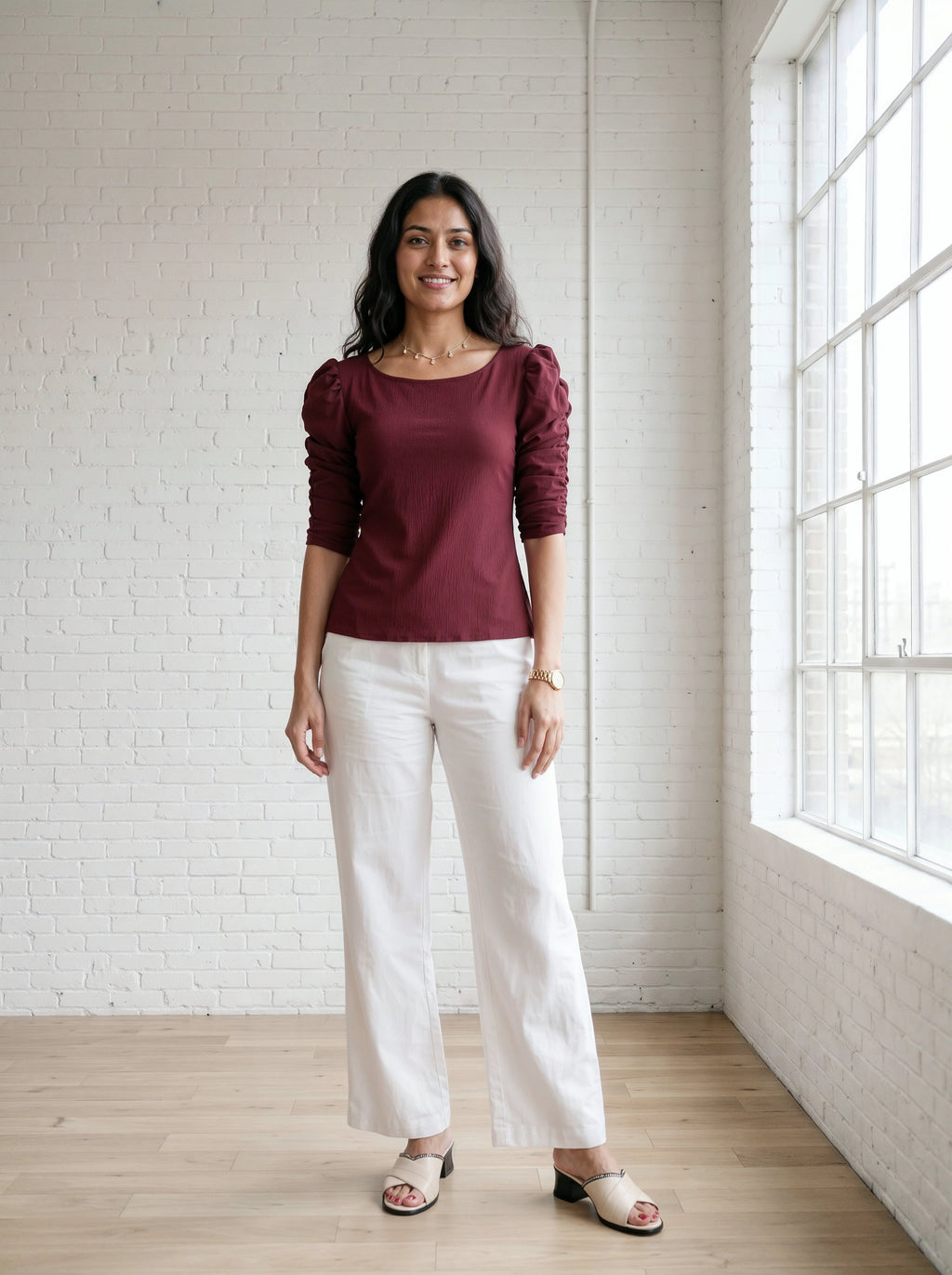 Woman wearing a burgundy top and white pants standing in a room with a white brick wall and large window.