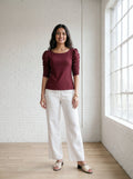 Woman wearing a burgundy top and white pants standing in a room with a white brick wall and large window.