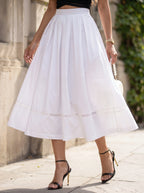 A woman wearing a dusty pink tiered midi dress with three-quarter sleeves and a buttoned collar, standing on a cobblestone patio in front of a beige building with outdoor cafe seating.
