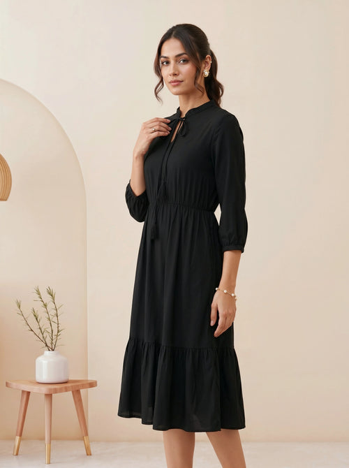 Woman wearing a black dress standing in a minimalistic room with a beige wall and wooden side table.