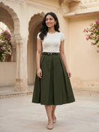 Woman wearing a white top and green skirt standing in front of an architectural background with flowers.