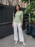 Woman in a green top and white pants standing outdoors with plants and a building in the background