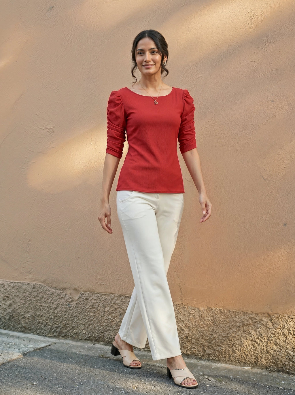 Woman wearing a red top and white pants standing against a beige wall.