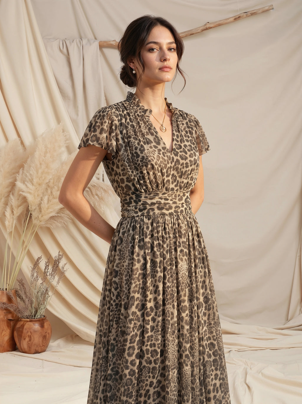 A woman wearing a leopard-print midi dress with short flutter sleeves, a V-neckline, and a gathered waist, posed against a draped beige fabric backdrop with dried pampas grass and wooden decor.