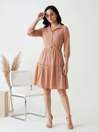 Woman wearing a peach-colored dress standing in a minimalistic room with a chair and decorative elements.