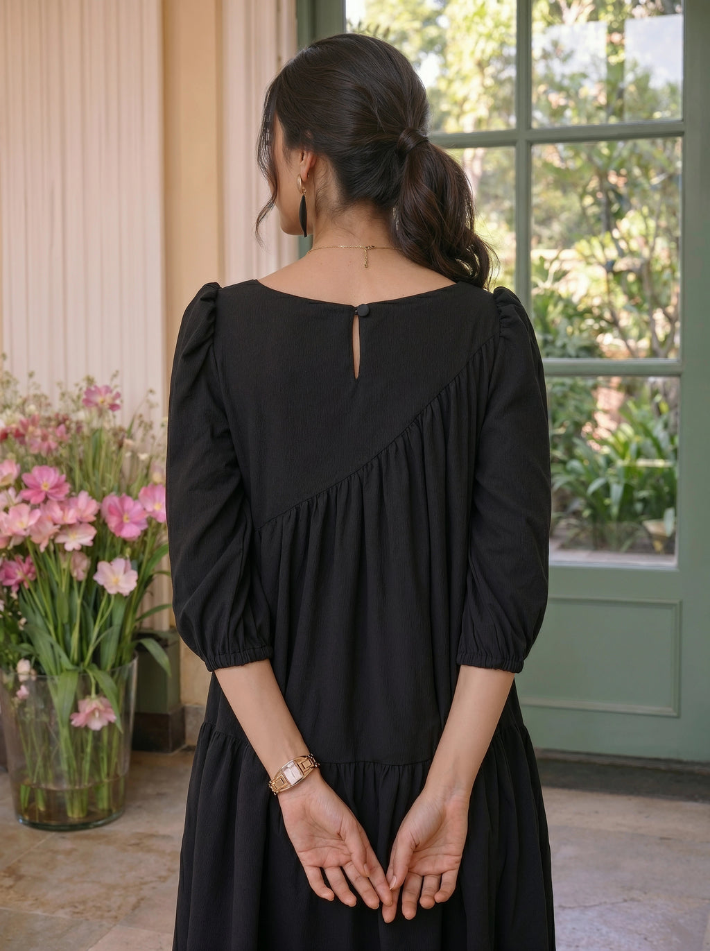 Woman wearing a black dress standing in a room with flowers and a window.