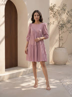 Woman in a pink dress standing in a sunlit outdoor setting with a wooden door and plant in the background.