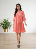 Woman wearing a coral dress standing in a room with a plant and white wall.