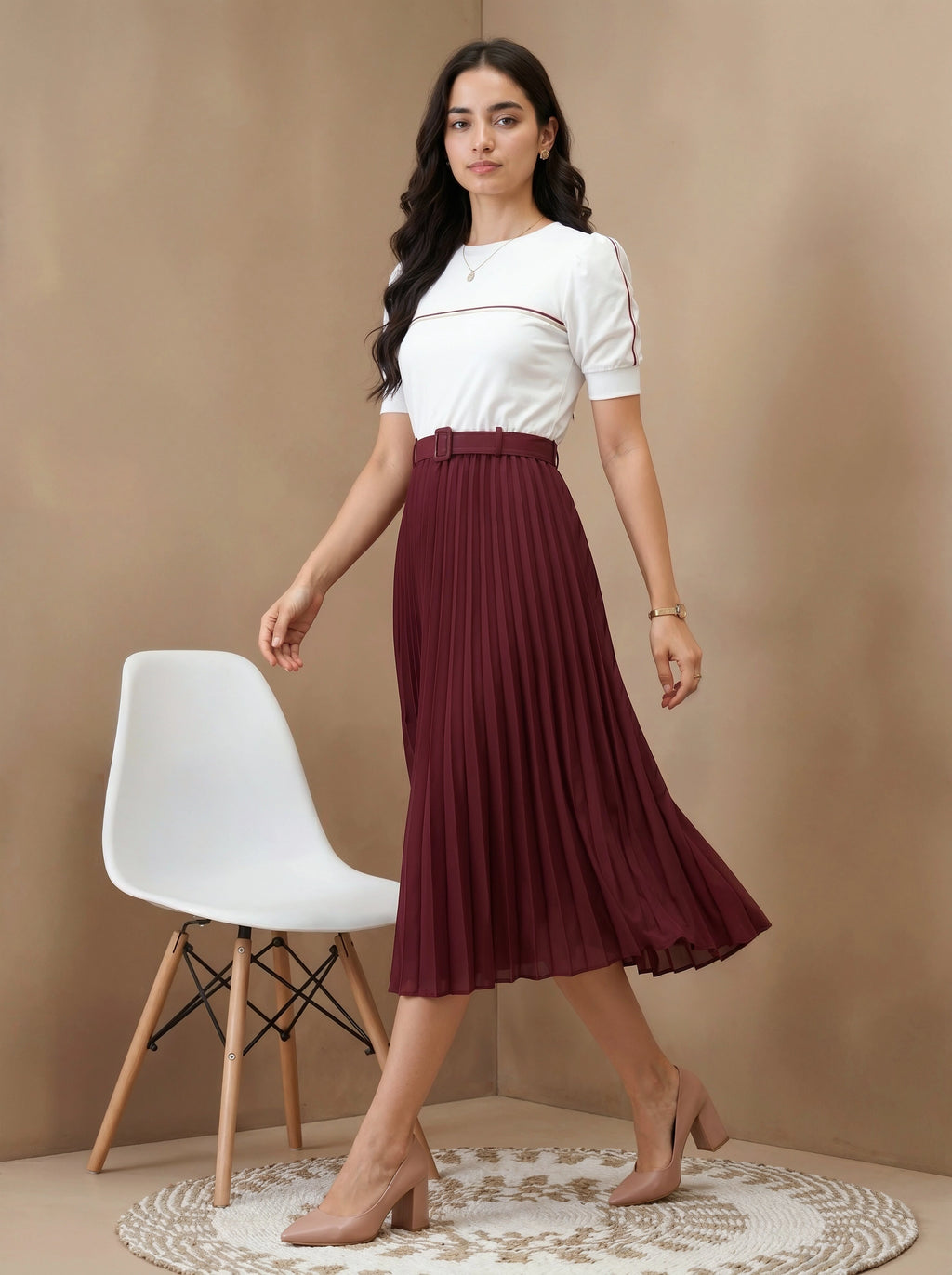Woman wearing a white top and burgundy pleated skirt standing in a room with a white chair and beige wall.