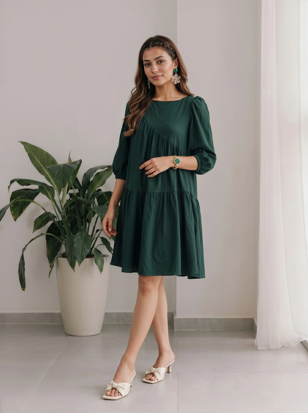 Woman wearing a green dress standing indoors with a plant and curtain in the background