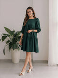 Woman wearing a green dress standing indoors with a plant and curtain in the background