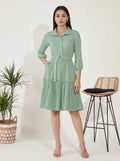 Woman wearing a green dress standing next to a plant and stool on a white background