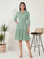 Woman wearing a green dress standing next to a plant and stool on a white background