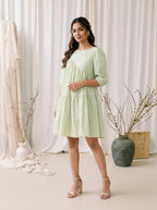 Woman wearing a light green dress standing in a softly lit room with white curtains and decorative vases.