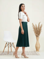 Woman wearing a white top and green skirt standing in a minimalistic room with a chair and vase.
