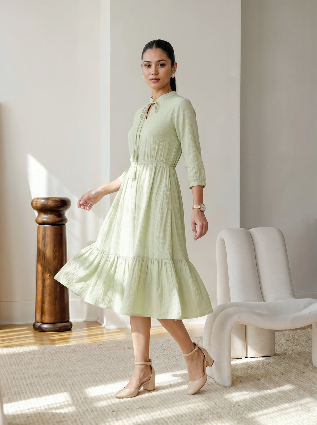 Woman in a light green dress standing in a bright room with a white chair and wooden column.
