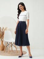 Woman wearing a white top and navy skirt standing in a minimalistic room with a chair and decorative items.