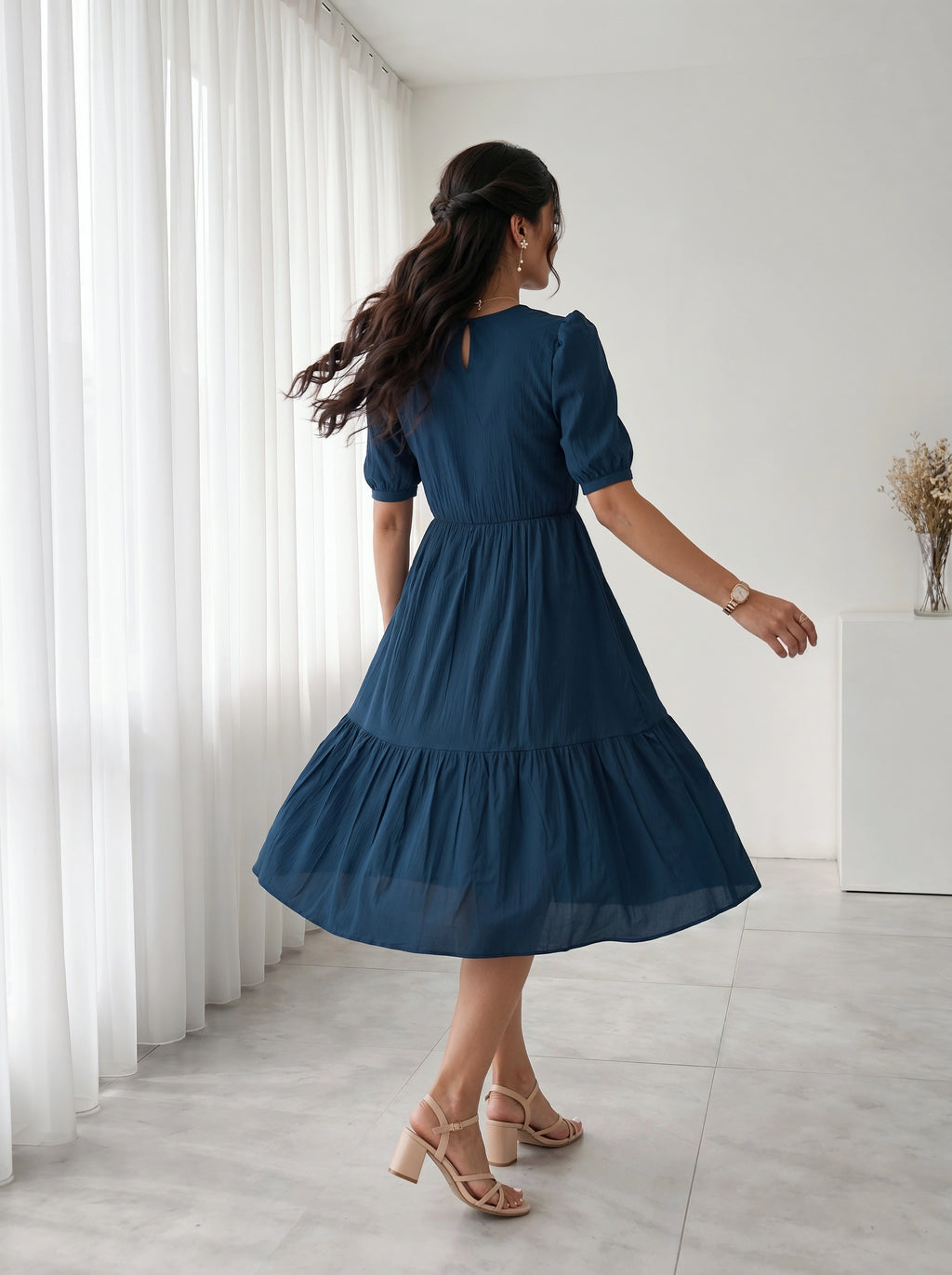 Woman in a blue dress standing in a room with white curtains and a white cabinet.