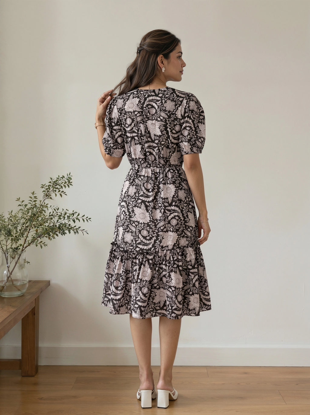 A woman stands with her back to the camera wearing a short-sleeved midi dress with puff sleeves and a tiered ruffle hem. The dress features a black and dusty pink floral pattern.