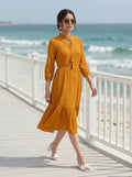 Woman in a mustard yellow dress standing on a wooden deck by the ocean.