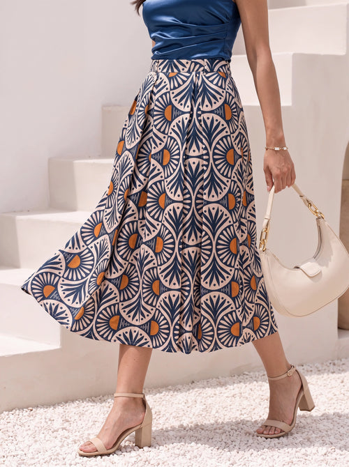 A woman wearing a high-waisted midi skirt with a navy blue, beige, and orange geometric fan pattern, paired with a blue top, beige heeled sandals, and carrying a cream-colored handbag.