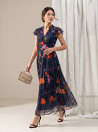 A woman models a navy blue maxi dress with a vibrant multicolor floral print, holding a beige handbag and wearing black high heels in a studio setting with white curtains.