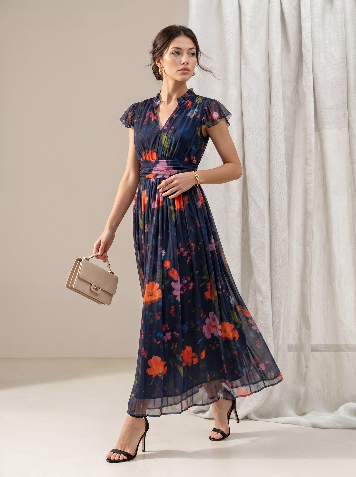 A woman models a navy blue maxi dress with a vibrant multicolor floral print, holding a beige handbag and wearing black high heels in a studio setting with white curtains.