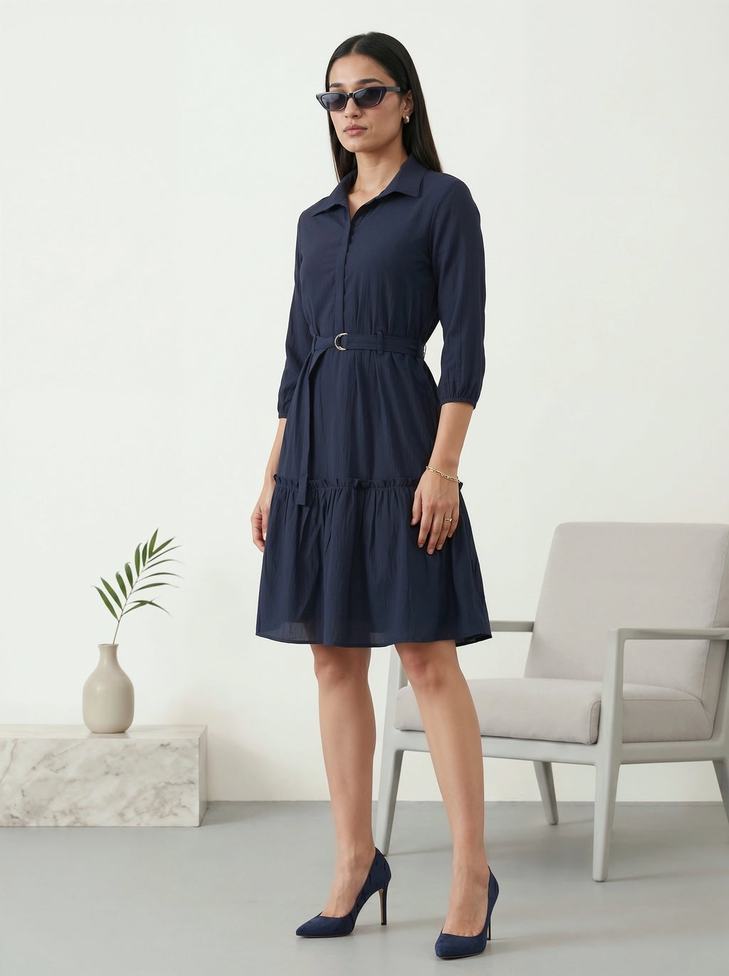 Woman wearing a navy blue dress standing in a minimalistic room with a chair and plant.