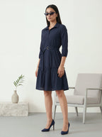 Woman wearing a navy blue dress standing in a minimalistic room with a chair and plant.