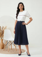 Woman wearing a white top and navy pleated skirt in a minimalistic room.