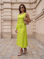 A woman wearing a bright lime green long-sleeve midi dress, black strappy high heels, and a beige shoulder bag, standing on a stone paved walkway in front of a beige stone building.
