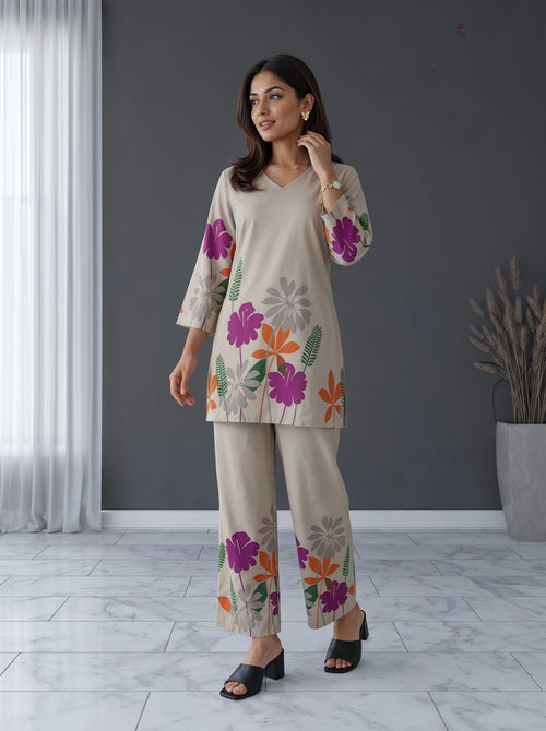 A woman models a two-piece beige pajama set with a colorful floral print, consisting of a V-neck long-sleeve top and matching wide-leg pants, in a modern room setting.
