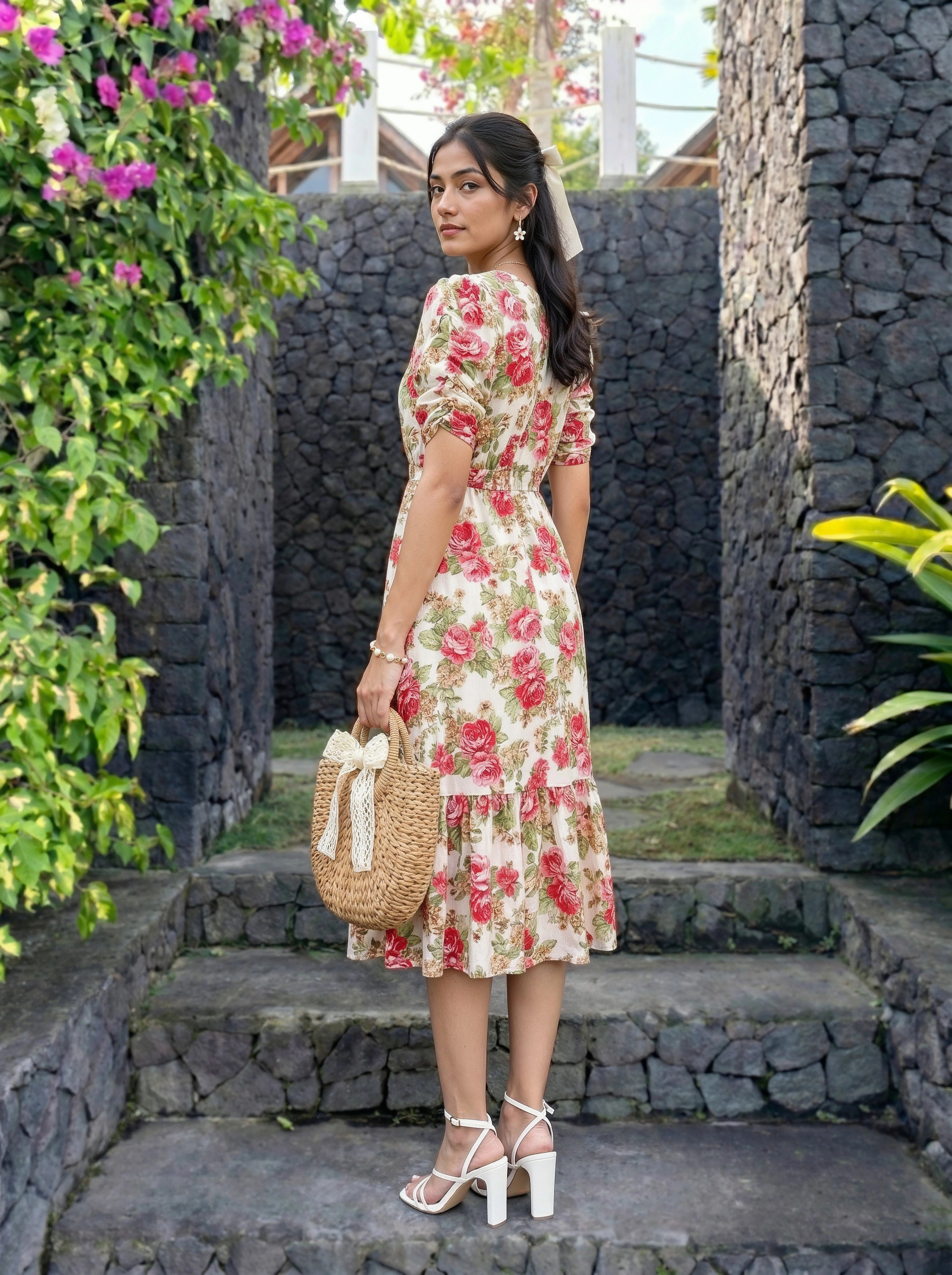 A woman stands on stone steps wearing a cream midi dress with a red and pink floral print, holding a woven straw bag with a white bow. The background features dark stone walls and lush green foliage with pink flowers.