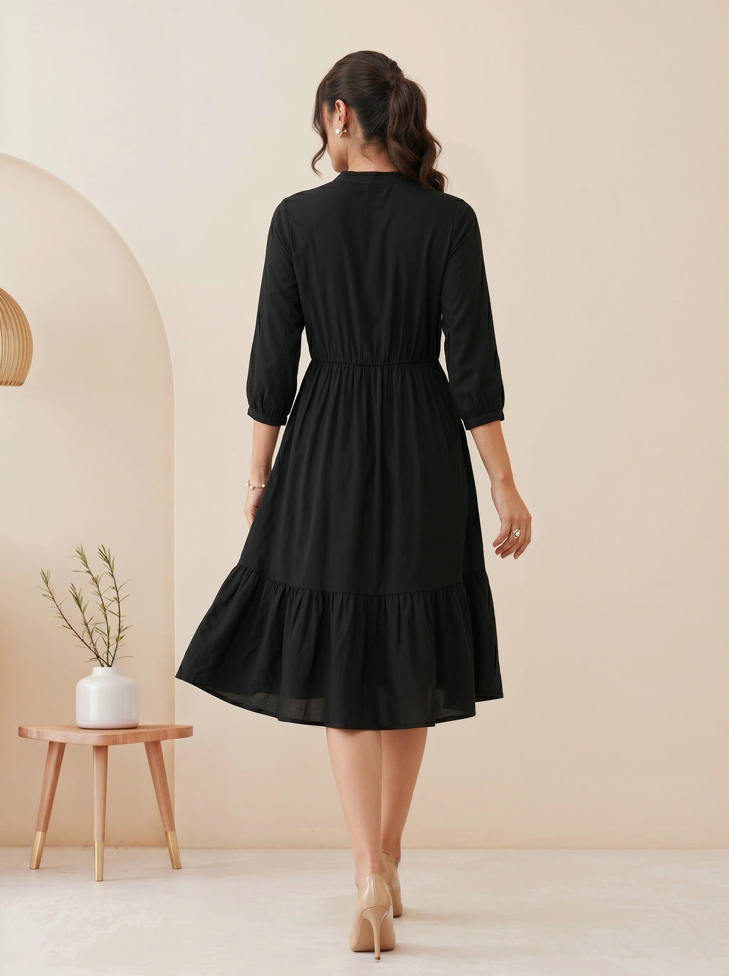 Woman wearing a black dress standing in a minimalistic room with a beige wall and small table.