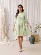 Woman in a light green dress standing in a softly lit room with white curtains and decorative vases.