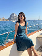 A woman wearing a sleeveless teal blue dress and sunglasses sits on the deck of a yacht, with the blue sea, rocky island, and boats in the background.