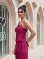 A woman modeling a magenta two-piece set consisting of a sleeveless camisole top with lace-up detailing and matching wide-leg trousers, set against a background of beige brick arches.