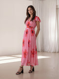 A woman modeling a pink midi dress with a red and black floral print, standing in a bright room with white curtains.
