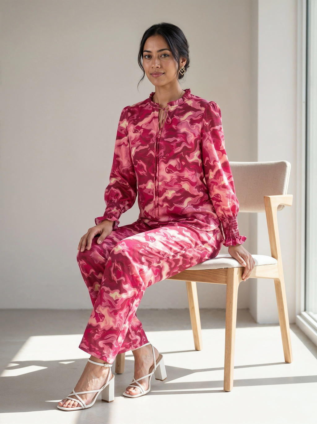A woman sits on a light wood chair wearing a matching pink and red camouflage print long-sleeve top and trousers set, paired with white heeled sandals in a bright indoor setting.