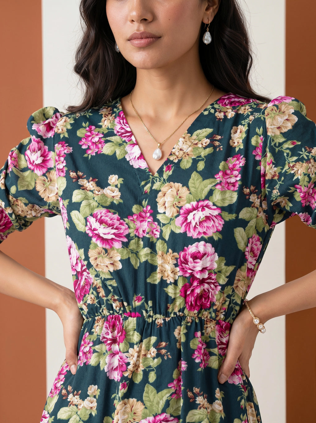A woman wearing a dark teal floral dress with pink, green, and beige flowers, featuring puff sleeves and a gathered waist, set against a split brown and white background.
