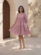 Woman in a pink dress standing in a sunlit outdoor setting with a wooden door and potted plant in the background.